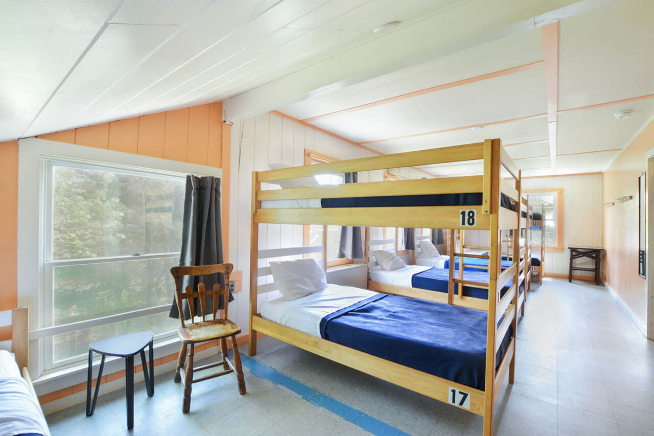 A large dorm room at HI Martha's Vineyard hostel with several sets of twin-sized bunk beds. There are several windows letting in natural light with views of the trees outside.
