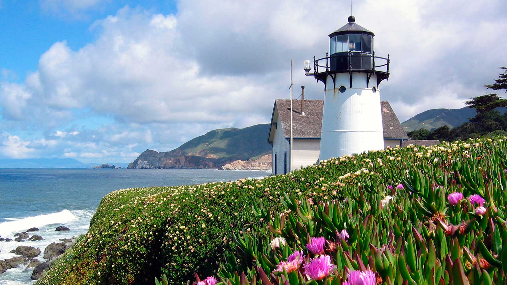 Point Montara Lighthouse, a historic white lighthouse set in front of the Pacific ocean with green ice plant and pink flowers in the foreground.
