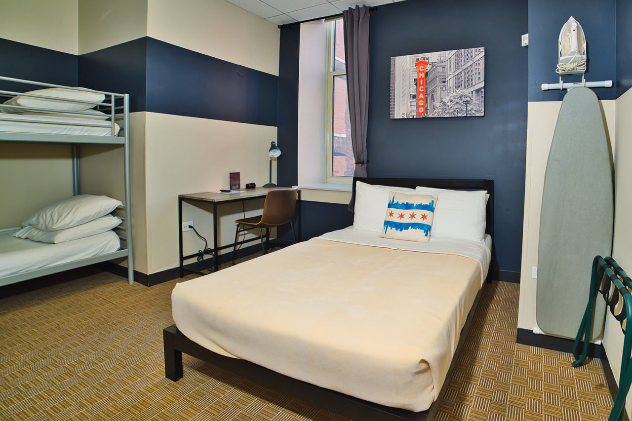 a private room at HI Chicago hostel sleeping up to three people. There is one freshly made full-sized bed and one set of twin-sized bunkbeds with clean sheets and pillows. An ironing board, luggage rack, and small desk with chair are also in the room.