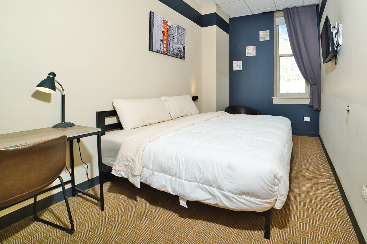 a private room with one freshly made king-sized bed at hi chicago hostel. There is a writing desk with lamp and a chair in the corner, and a large window with open curtains letting in natural light.