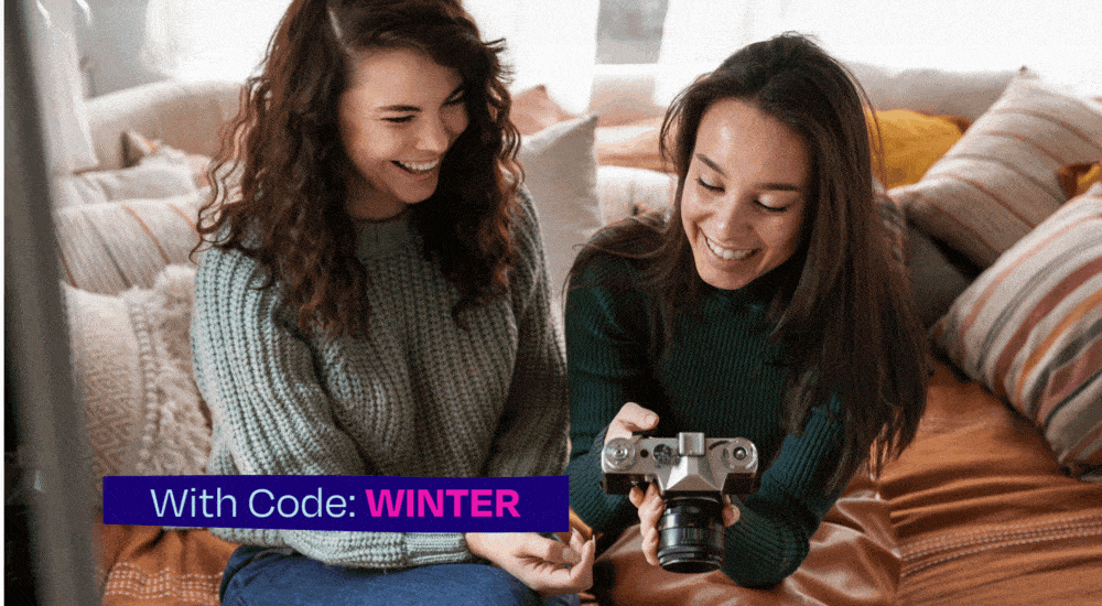 Two young travelers in sweaters sit on a cozy couch surrounded by pillows looking at photos on a large black camera.