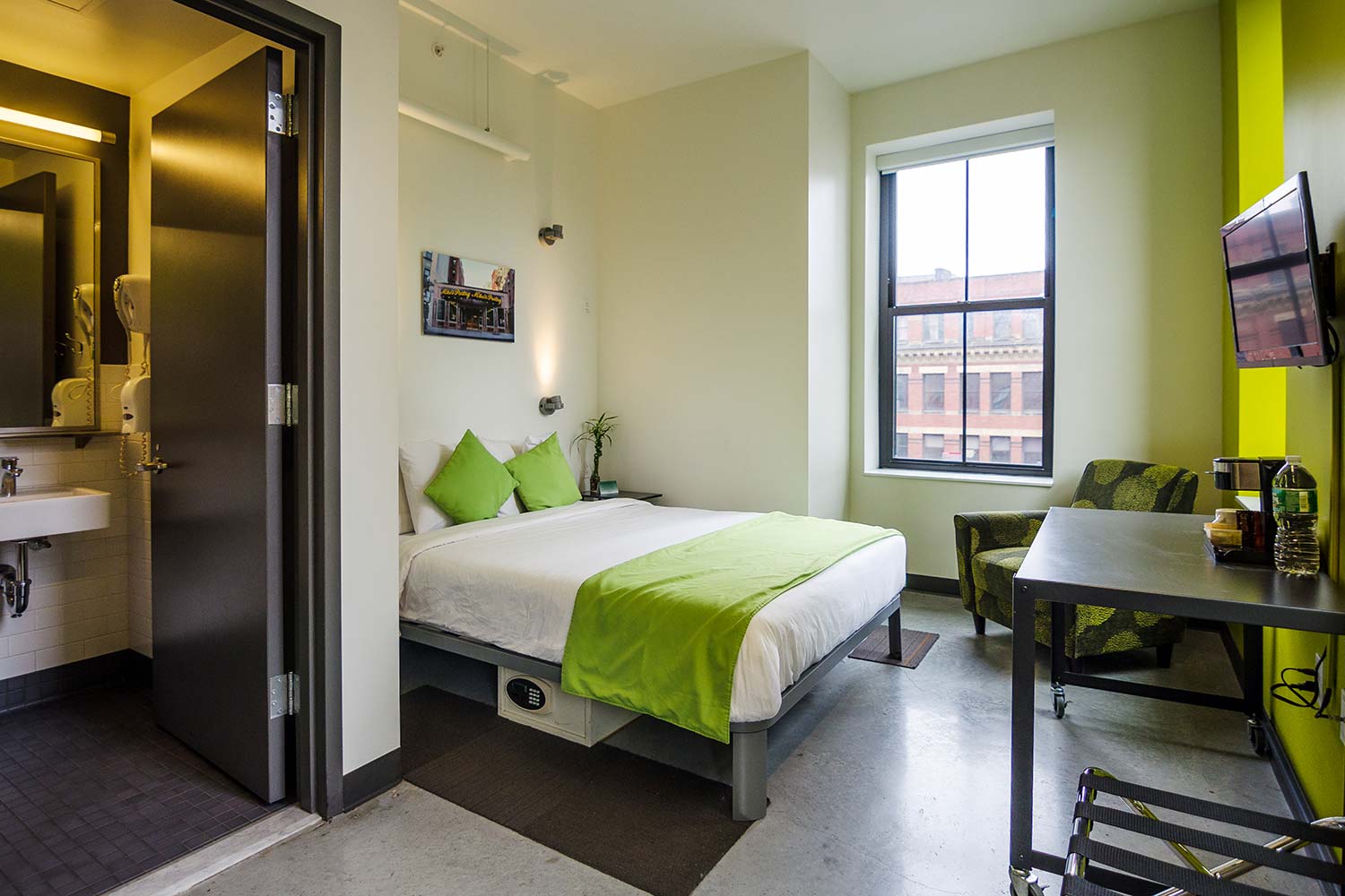 a private room at HI Boston hostel sleeps up to two people in a queen-sized bed. Opposite the bed, there is a small desk and a wall-mounted TV. Through an open door inside the room, a private ensuite bathroom is visible.