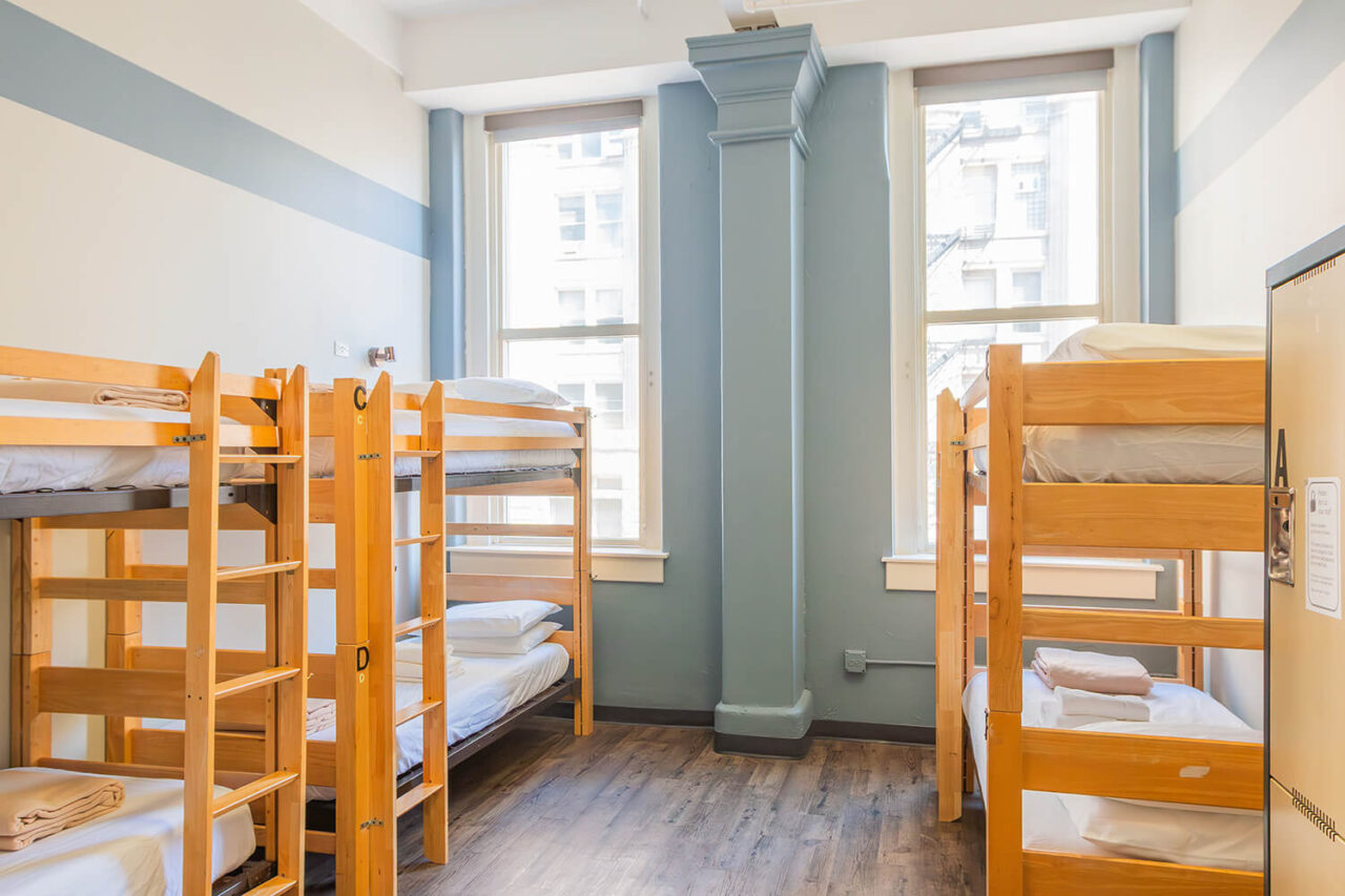 A dorm room at HI Chicago hostel with three sets of bunk beds. Each bed has fresh linens and an assigned locker. The walls are pale blue and there are a two large windows letting in natural light.