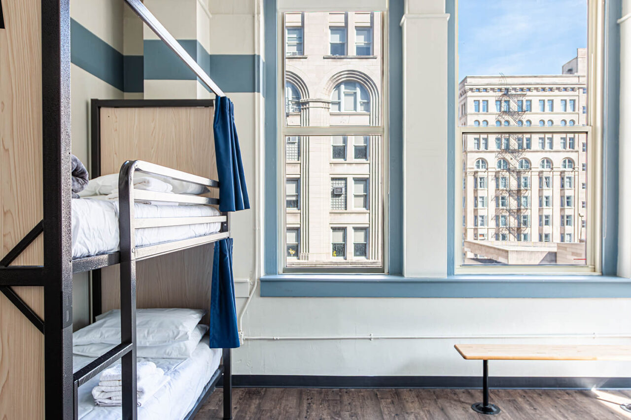 A spacious 4-bed dorm room at HI Chicago hostel showing bunkbeds with fresh linens and privacy curtains. On the wall are two large windows letting in plenty of natural light.
