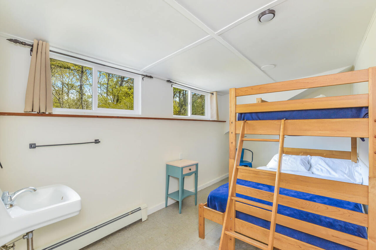 The four-person room with half ensuite bath at HI Martha's Vineyard hostel features one twin-sized bed bunked over one full-sized bed, one individual twin-sized bed, a toilet, and a shower, in a room with windows overlooking the trees.