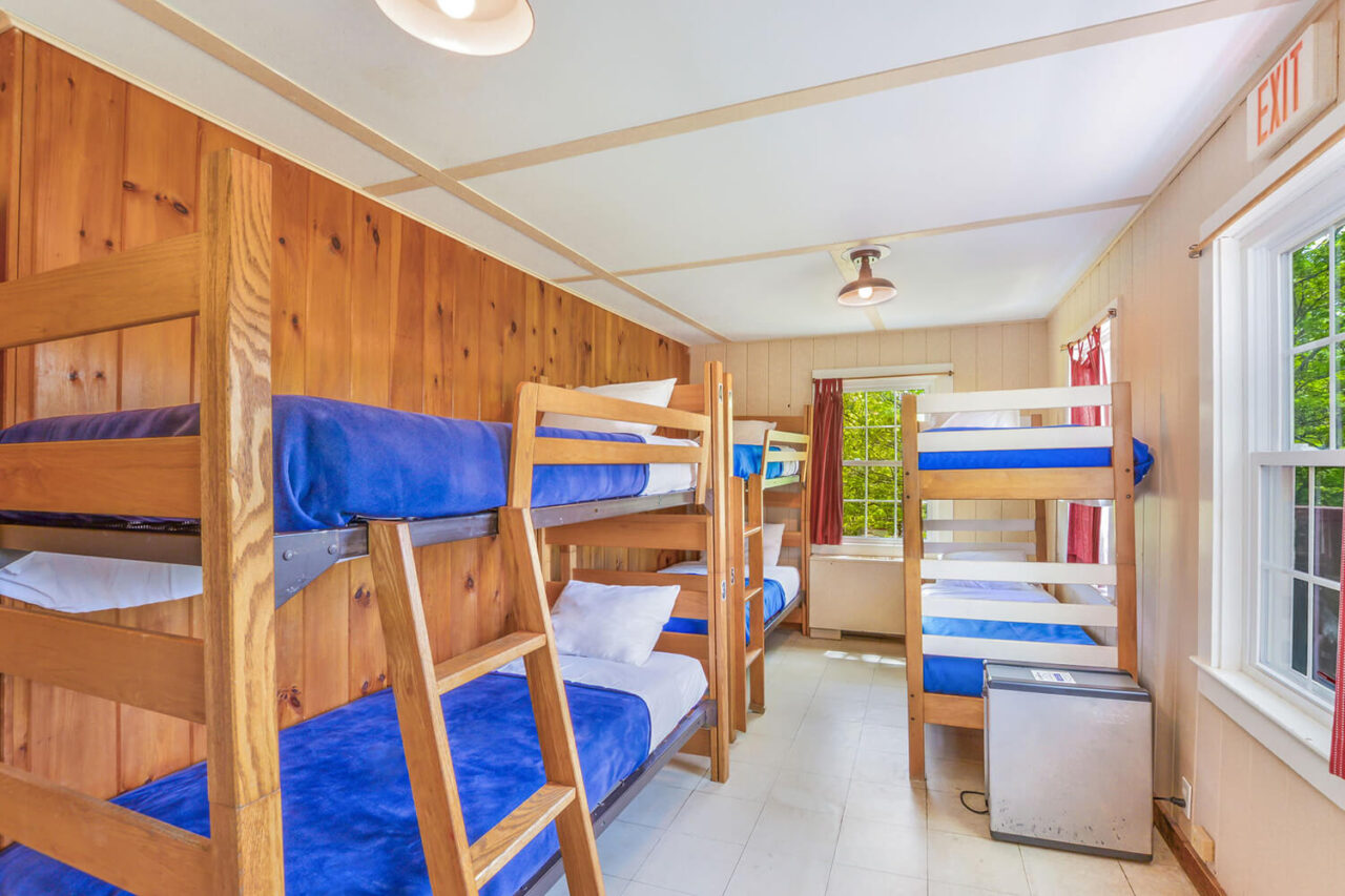 A dorm room at HI Martha's Vineyard hostel with four sets of twin-sized bunk beds and one individual twin-sized bed. There is wood paneling on the walls and a window overlooking the trees.