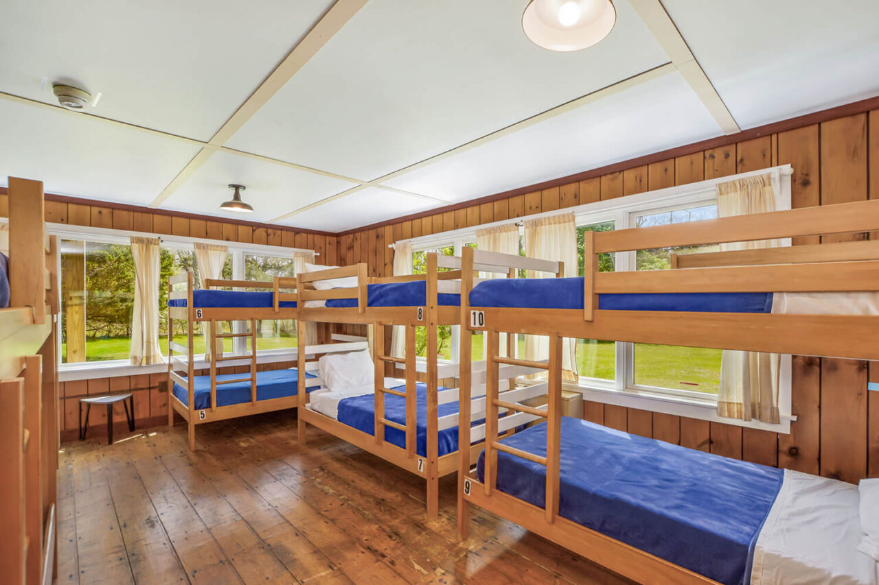 A bright and airy dorm room at HI Martha's Vineyard with five sets of twin-sized bunk beds, wood floors, and windows overlooking greenery.