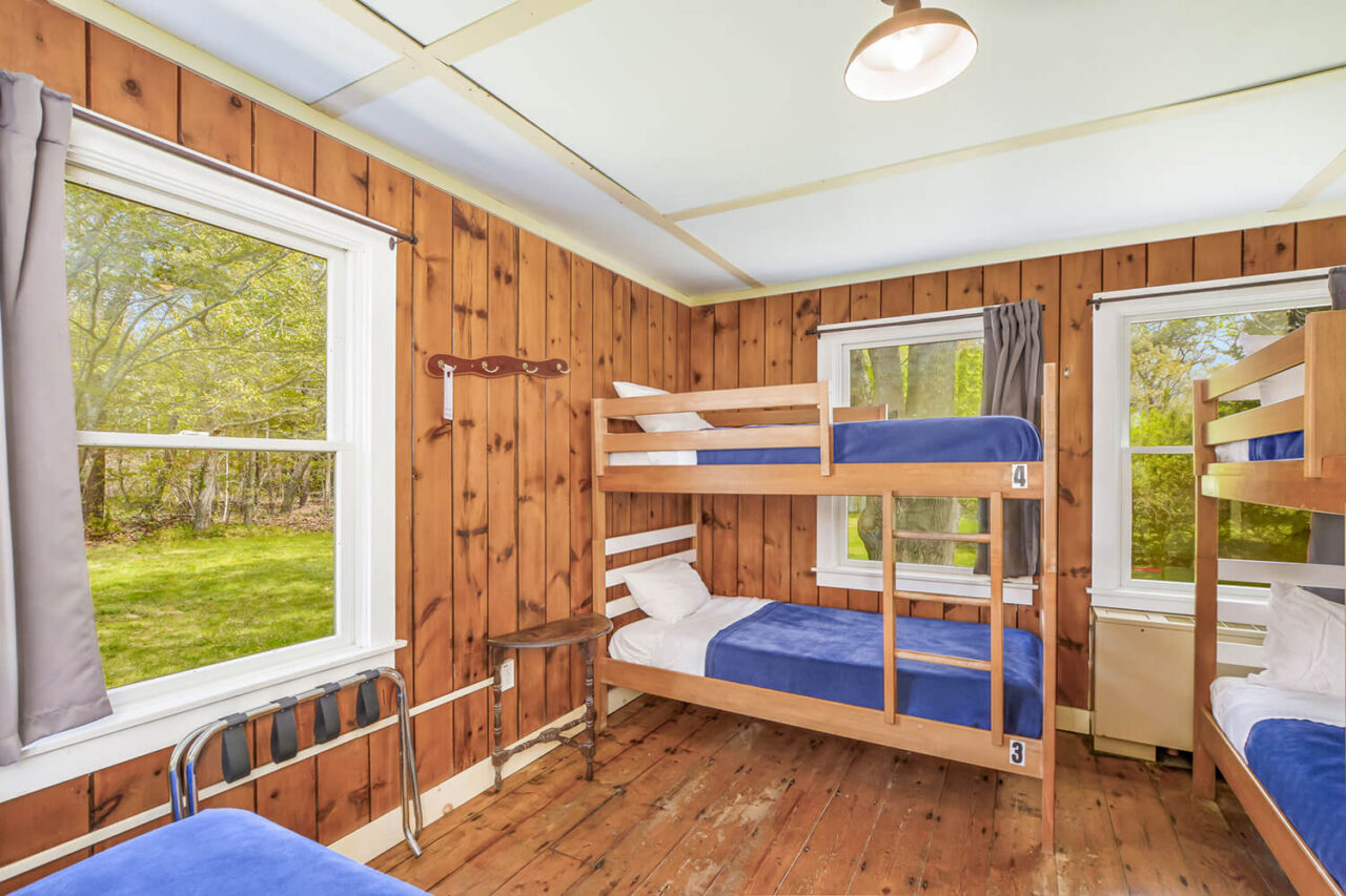 A large private room at HI Martha's Vineyard hostel contains one full-sized bed and two sets of twin-sized bunk beds. The walls are paneled in wood and there is a window overlooking the yard.