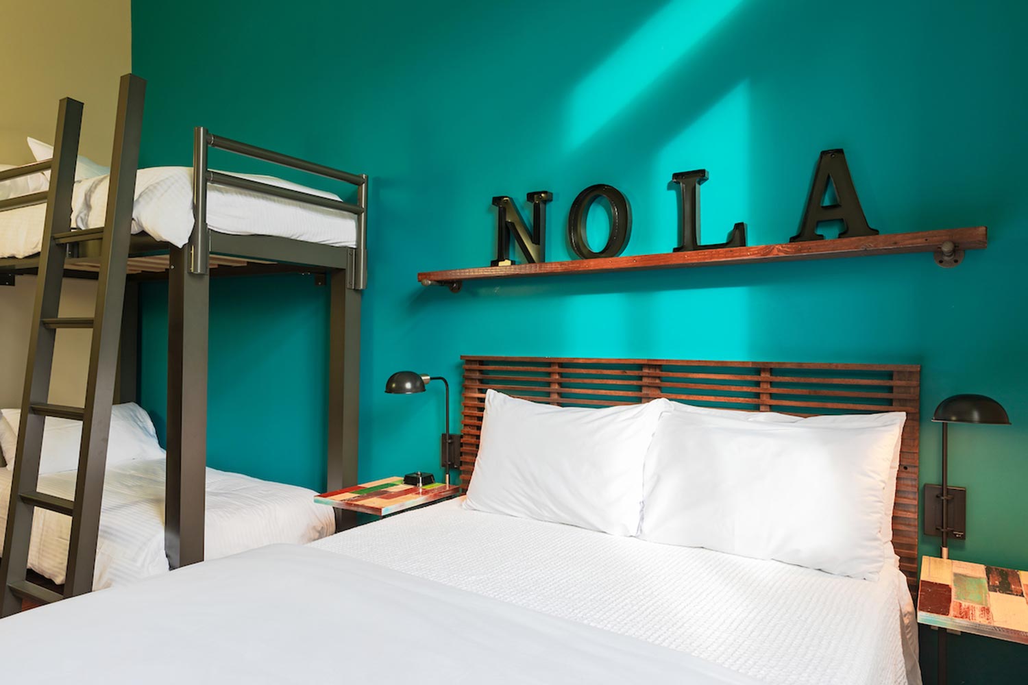 A private family room at HI New Orleans hostel with turquoise-colored walls and one freshly made full-sized bed with crisp white linens and one set of twin-sized bunk beds with crisp white linens.