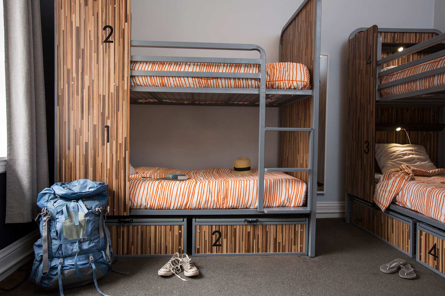 A dorm room at HI San Francisco Downtown hostel with four sturdy bunk beds with orange bedspreads. Each bed has secure built-in lockers below for guests' belongings. In front of one of the lower bunks, there is a pair of shoes and a large backpack.