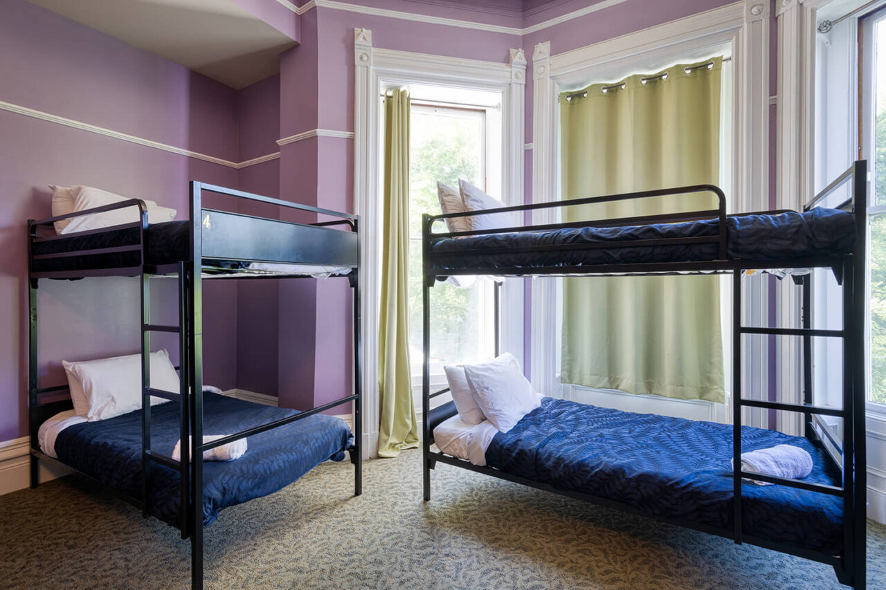 A shared dorm room at HI Sacramento hostel with a decorative fireplace, pale purple walls, large windows, and several sets of twin-sized bunks