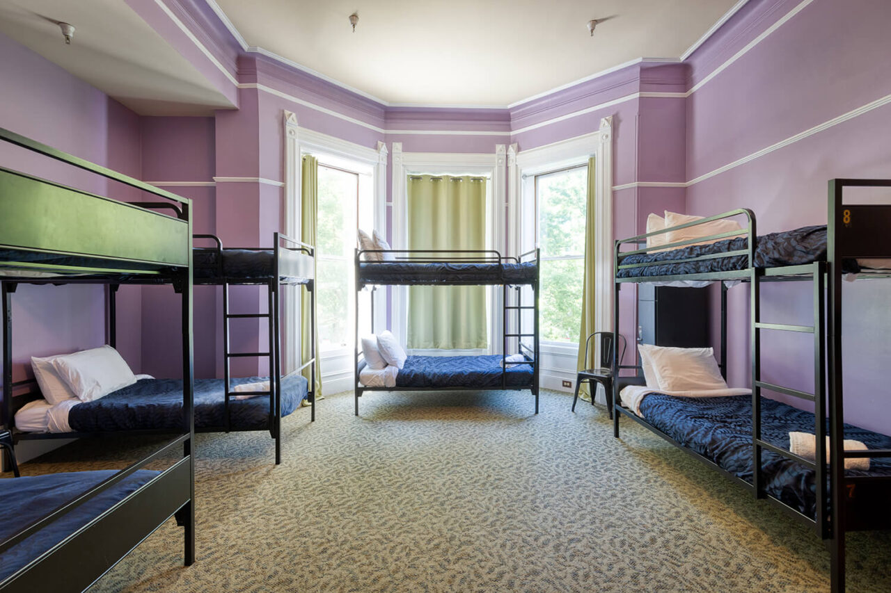 A shared dorm room at HI Sacramento hostel with a decorative fireplace, pale purple walls, large windows, and several sets of twin-sized bunks