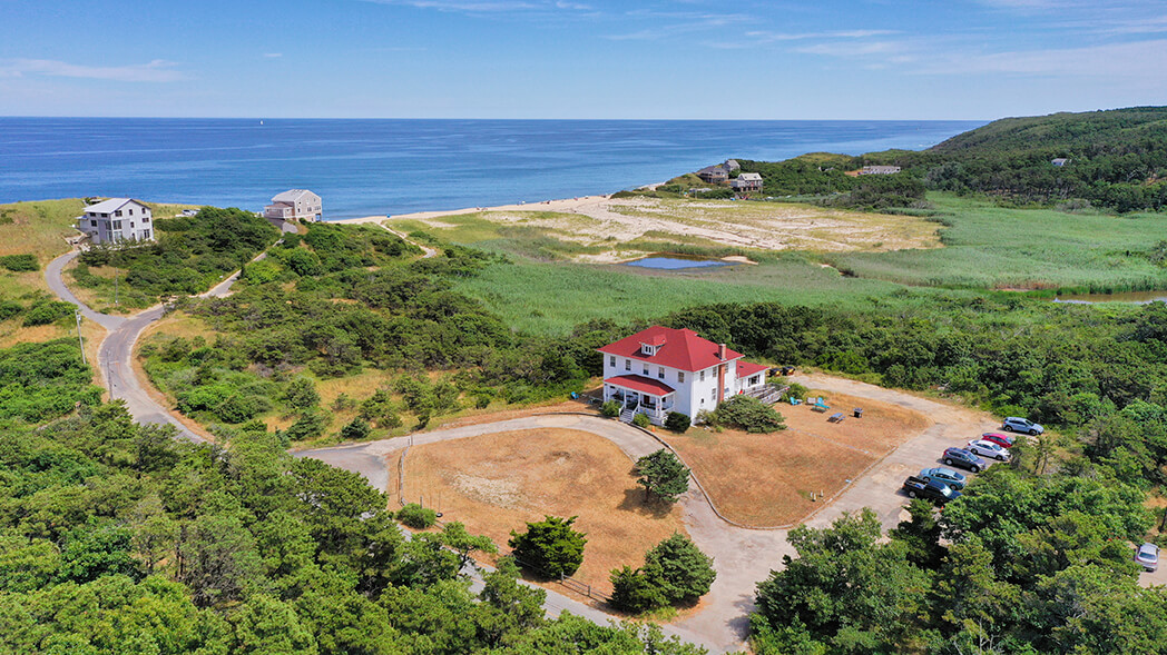 An aerial view of HI Truro hostel, a white building with a red roof, which sits surrounded by trees and looks out over nearby Ballston Beach