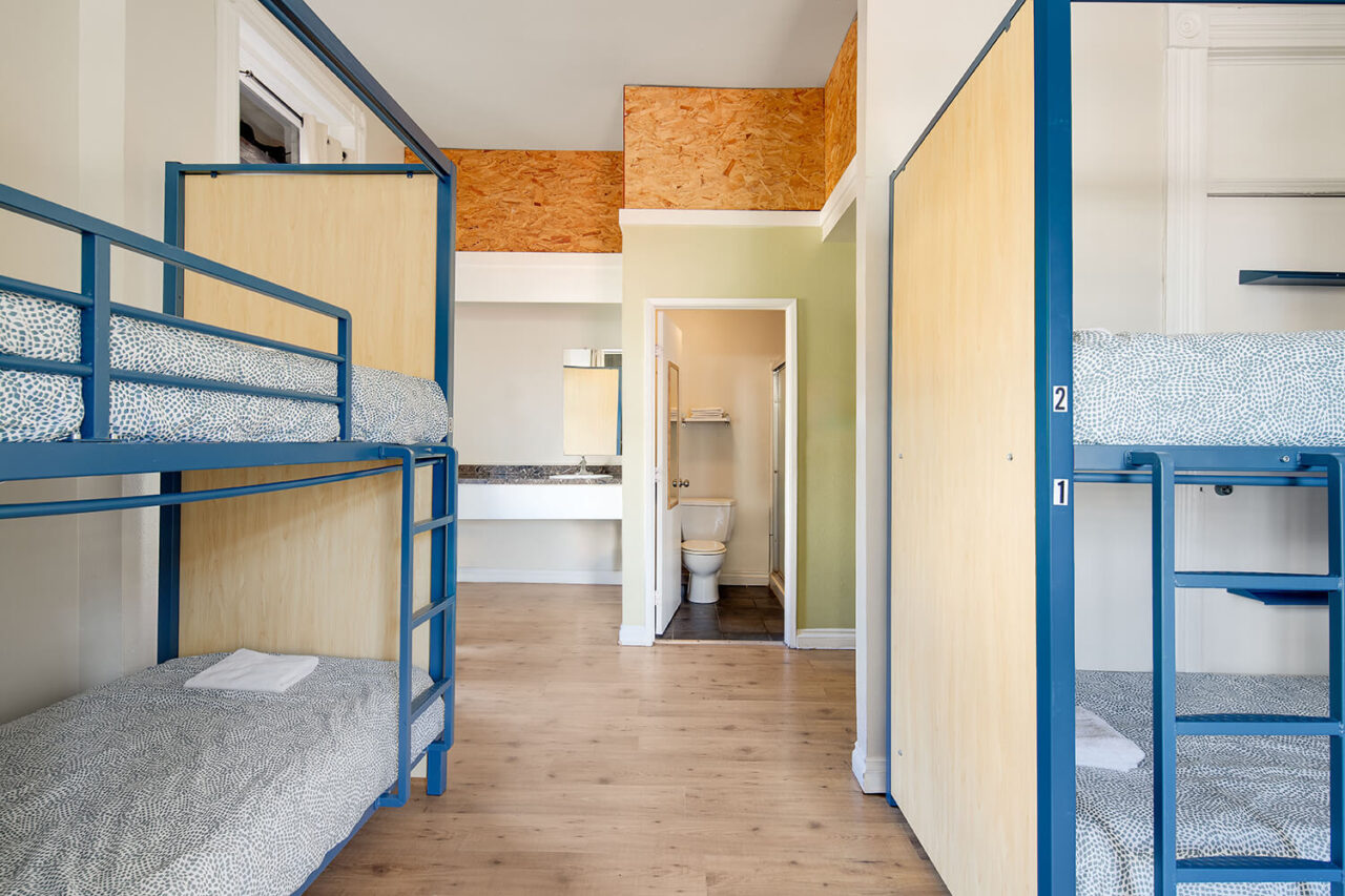 a sunlit dorm room at Hi San Diego Downtown hostel with privacy screens between bunkbeds and an ensuite bathroom visible in the background