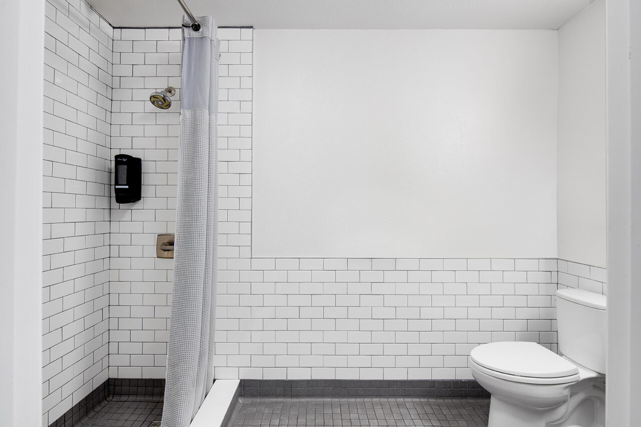a spacious single-occupancy shared bathroom at HI san Diiego Downtown hostel with a large standing shower, a toilet, and walls covered in clean white tile