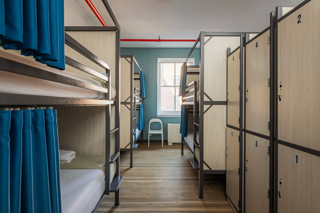 A modern shared dorm room at HI New York City hostel with 6 bunk beds with privacy screens and blue privacy curtains on each bed, individual lockers, wood floors, and a blue wall with window.