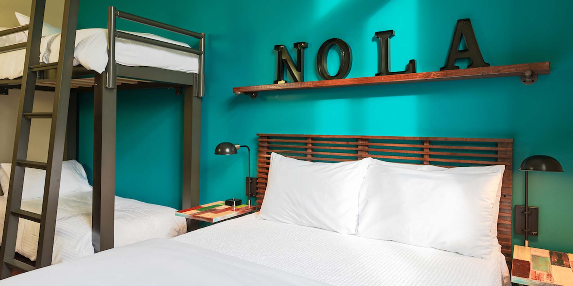 A private family room at HI New Orleans hostel with turquoise-colored walls and one freshly made full-sized bed with crisp white linens and one set of twin-sized bunk beds with crisp white linens.