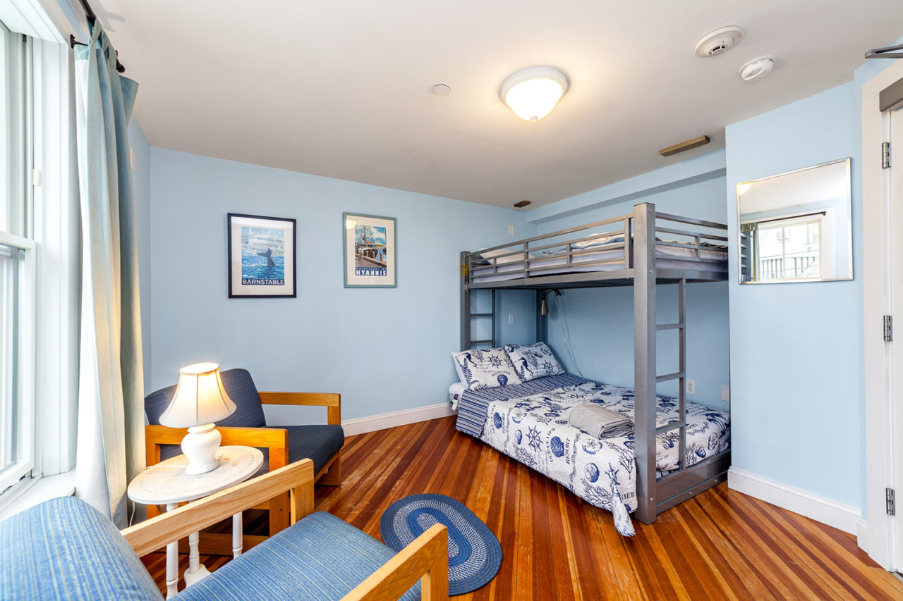 a spacious and airy room with wood floors, light blue walls, one set of freshly made bunk beds, and an armchair