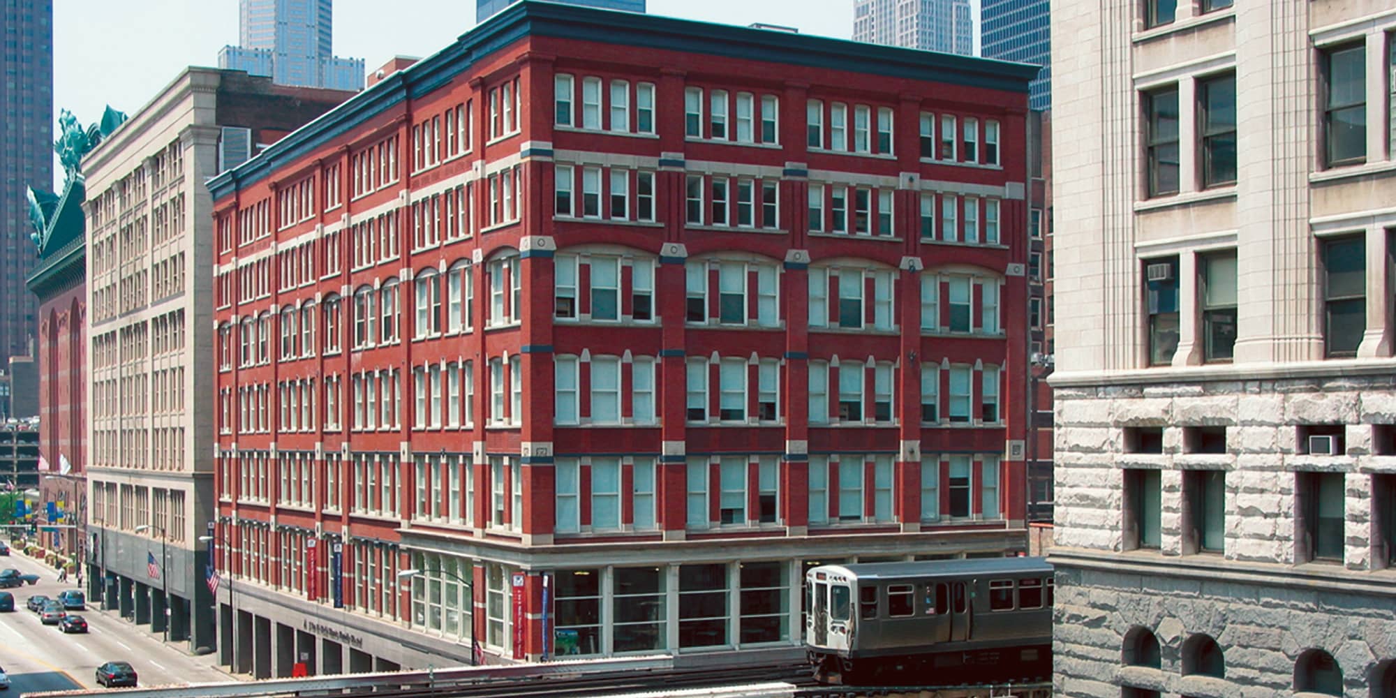 the exterior of HI Chicago hostel, a large brick building, as seen from the street. An L subway car runs on an elevated platform in the foreground.