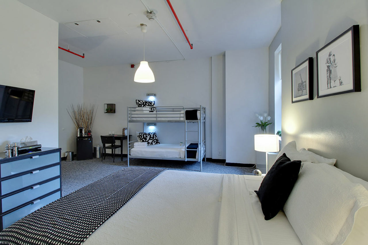 A large family private room at HI New York City hostel. There is one large bed facing a chest of dresser drawers with a TV mounted on the wall above it. On the other side of the room is one set of twin-sized bunk beds. All beds are made up with crisp white linens. The carpet is grey and the walls are white.