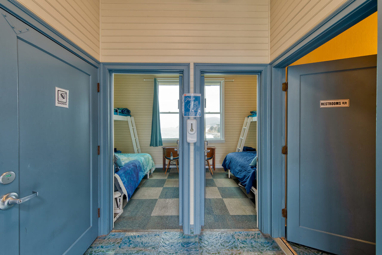 Through two open doors next to each other, a view inside of two separate rooms at HI Point Montara Lighthouse hostel. Each room in the two-room suite has one full-sized bed, one pullman-style twin bed, and a small desk and chair. The rooms share a wall, and each room has a window looking out over the coast.