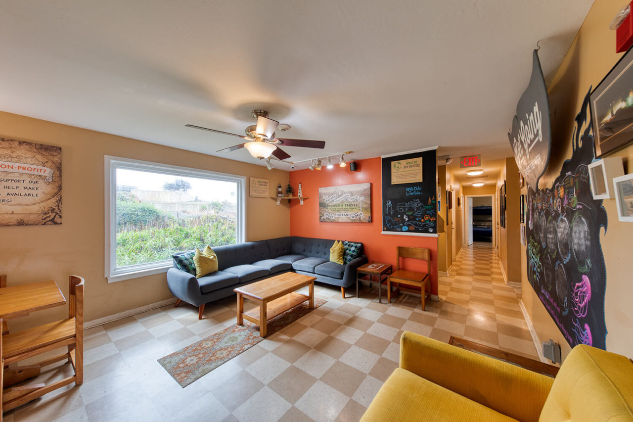 a large, brightly lit living room is available for guests of HI Point Montara Lighthouse hostel's Surfside House. The living room has a sectional sofa, armchair, and a view of the on-site garden.