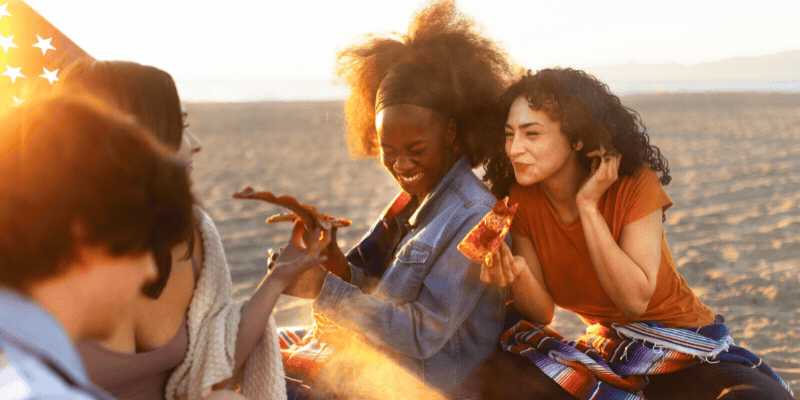 Two young girls eat pizza while laughing on the beach at sunset. Overlay text reads 