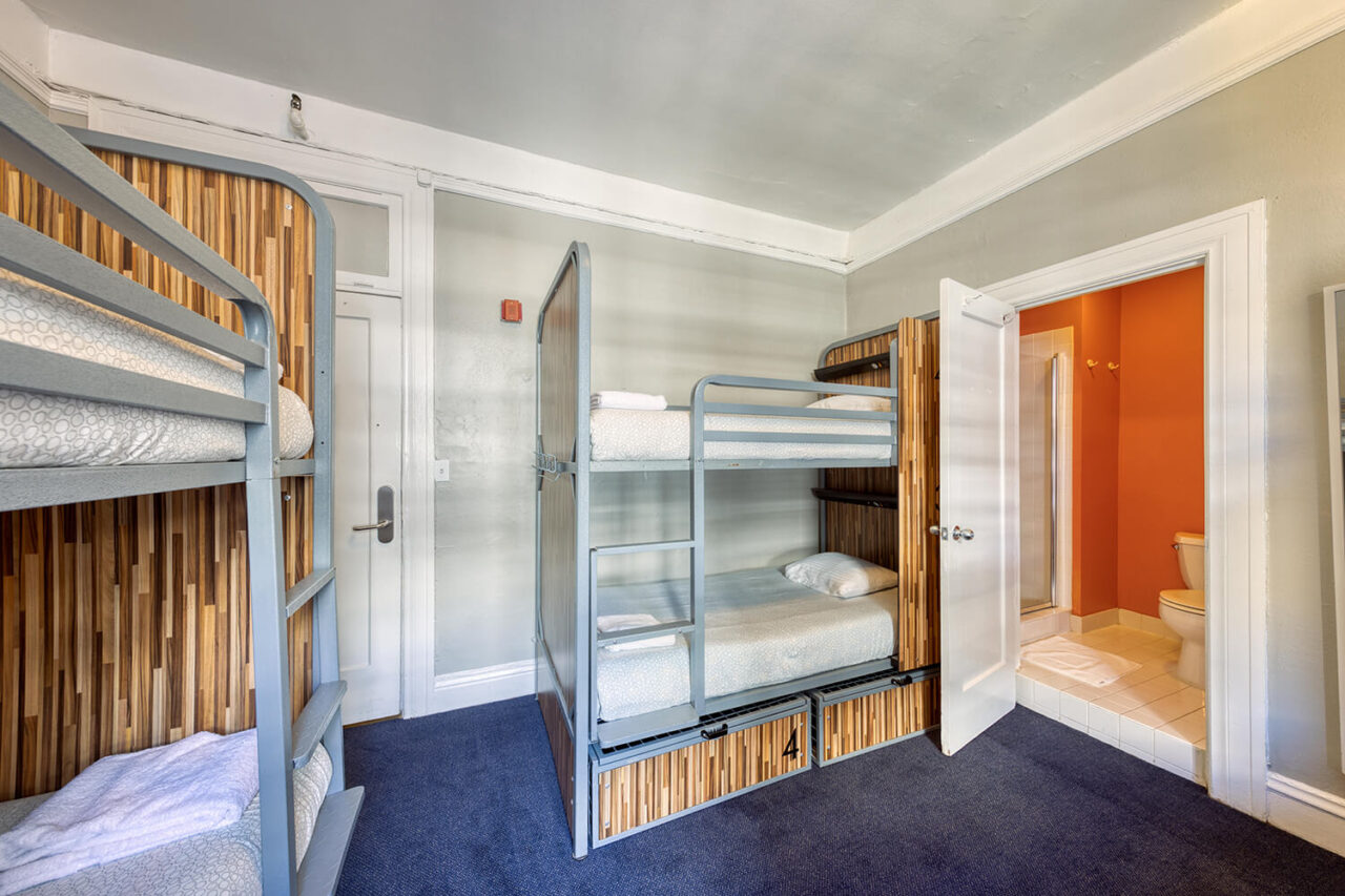 At HI San Francisco Downtown hostel, four modern bunk beds with privacy screens and build-in luggage storage. Next to one set of bunks, a door is open leading into a private bathroom