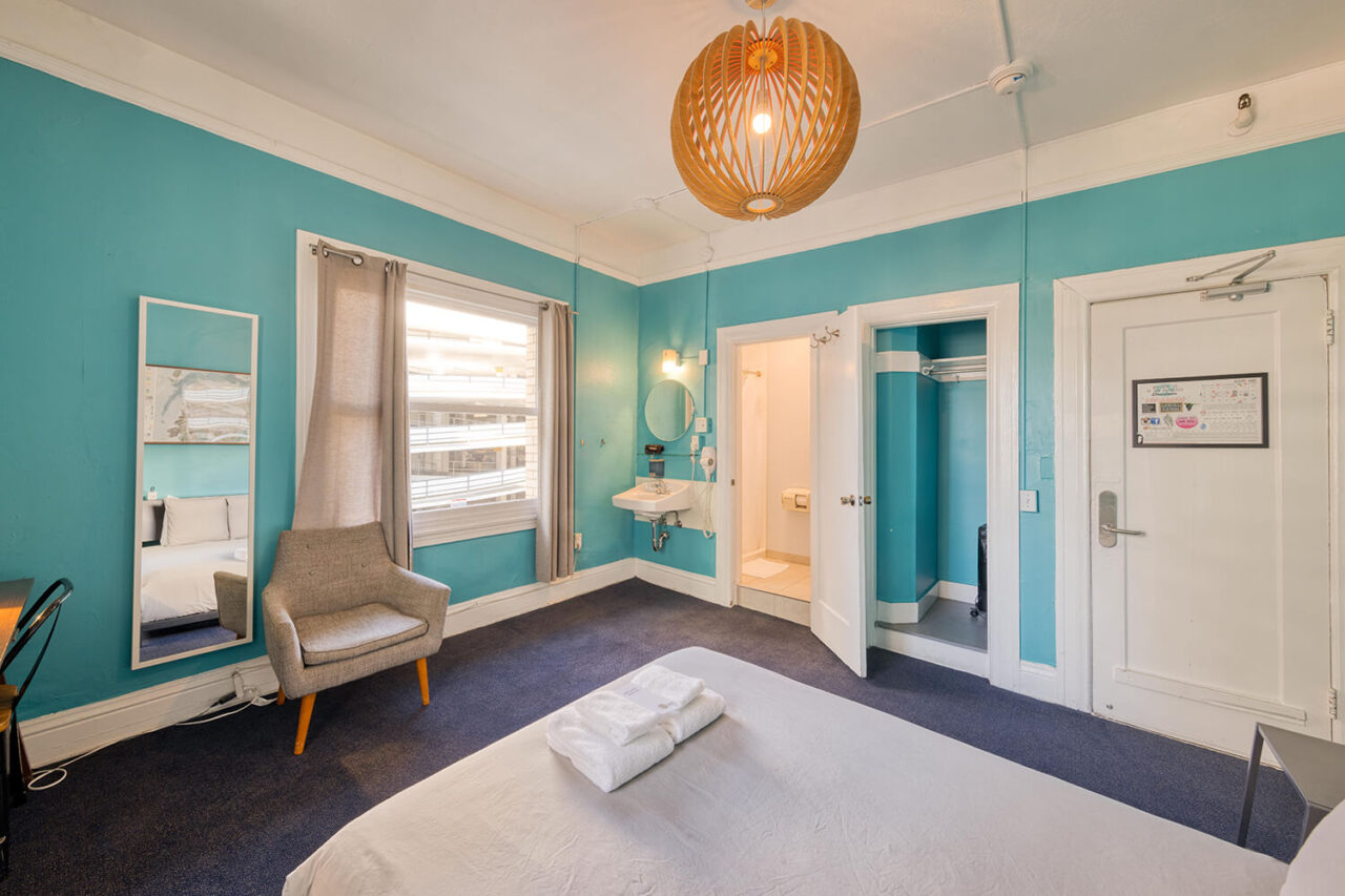 a queen-sized bed looks out across a private room with teal walls at HI San Francisco Downtown hostel. There is a grey arm chair in the room, and an internal door opens to show a private ensuite bathroom.