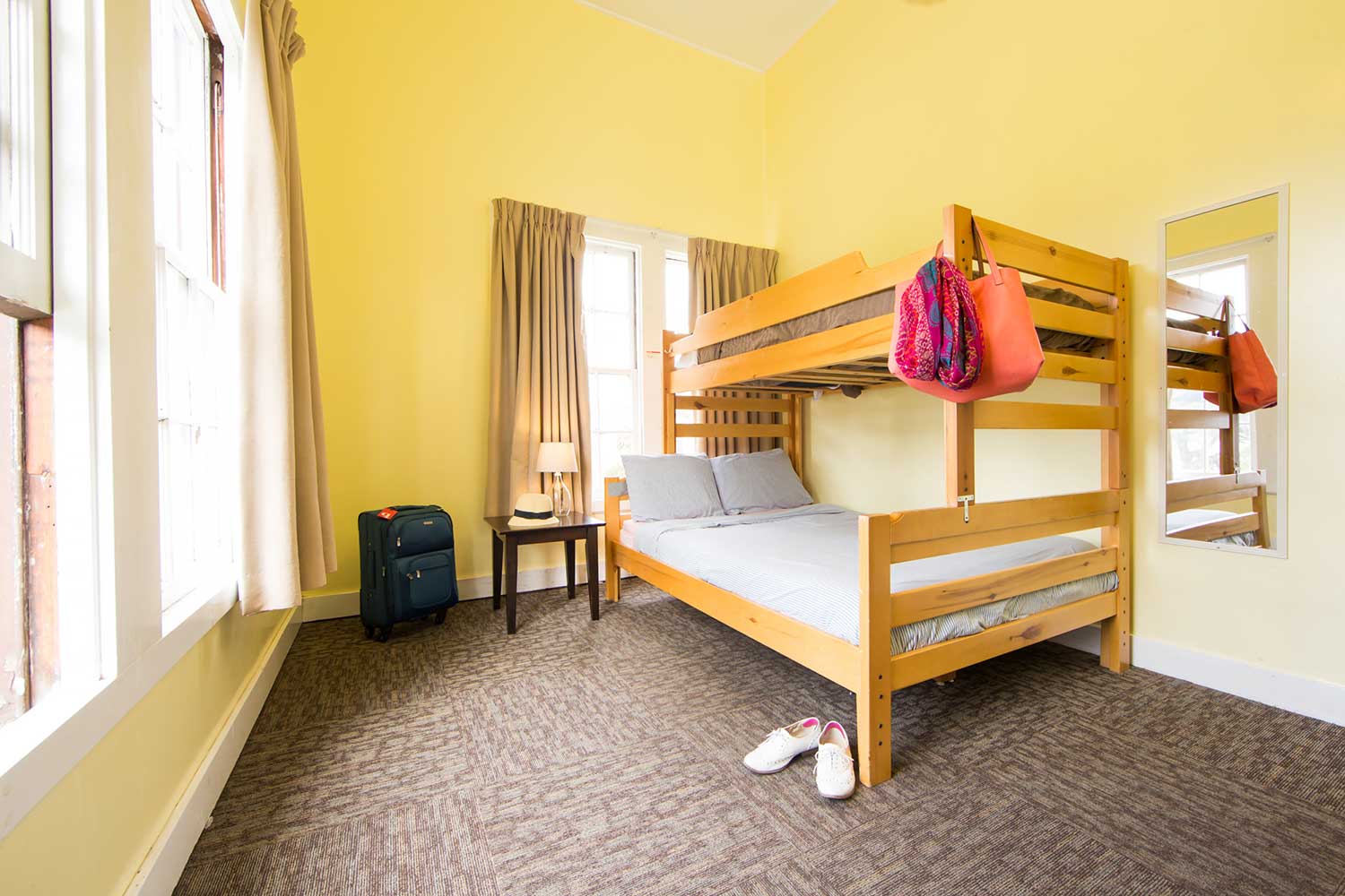 a private room at HI San Francisco Fisherman's Wharf hostel with one full-sized bed and one twin-sized bed bunked over it. The walls are a soft yellow color. There is a suitcase in the corner, and a pair of shoes and a bright pink scarf and handbag near the foot of the bed.