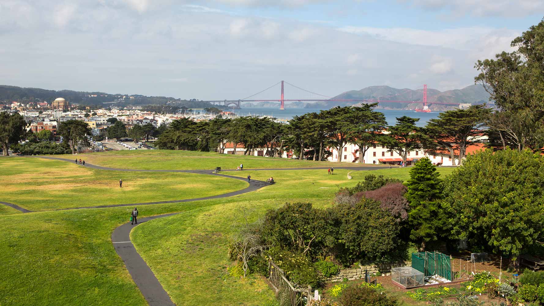 HI San Francisco Fisherman's Wharf hostel is located inside Fort Mason. An aerial view of the hostel shows the building's surroundings of grass, trees, and the nearby golden gate bridge spanning the San Francisco Bay.