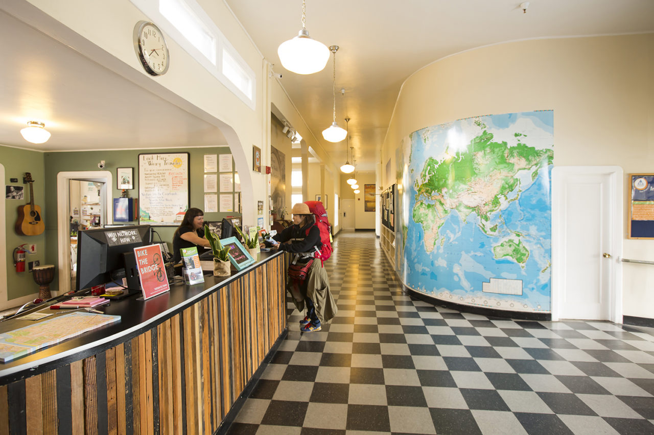 A guest checks in at the wood-paneled front desk of HI San Francisco Fisherman's Wharf hostel. There is a black-and-white checkered floor and a large map of the world on the wall.