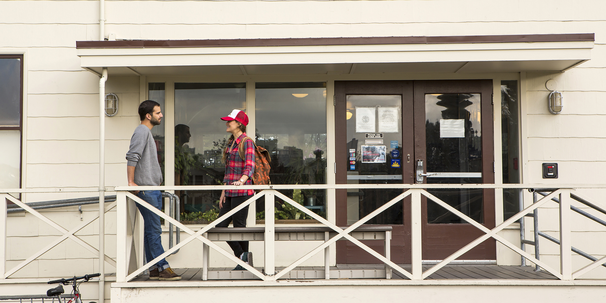 Two travelers stand talking at the entrance of HI San Francisco Fisherman's Wharf hostel. It is a historic building with a white exterior and an elevated, ADA-accessible entrance.