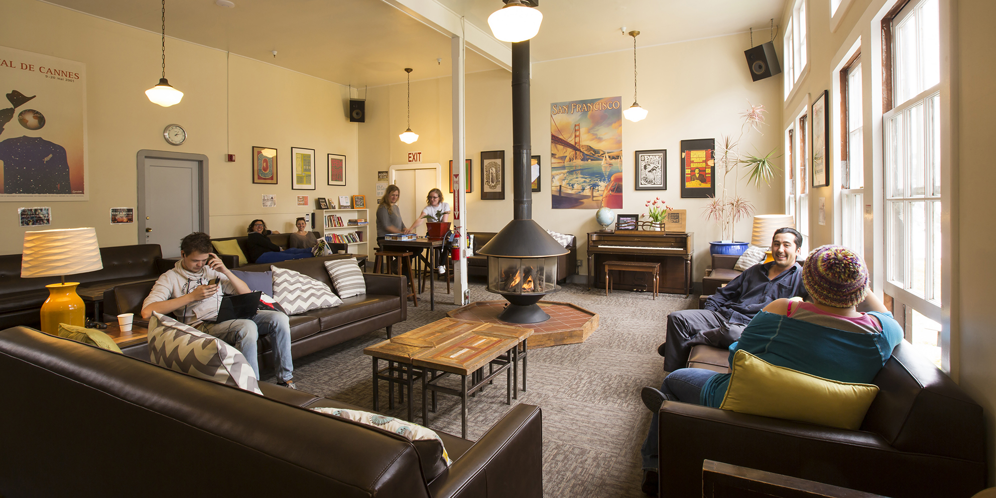 Guests gather in the lounge at HI San Francisco Fisherman's Wharf hostel. There is a fireplace in the middle of the room, a piano along the back wall, and multiple comfortable couches and armchairs where travelers are relaxing. On the white walls hang vintage concert posters.