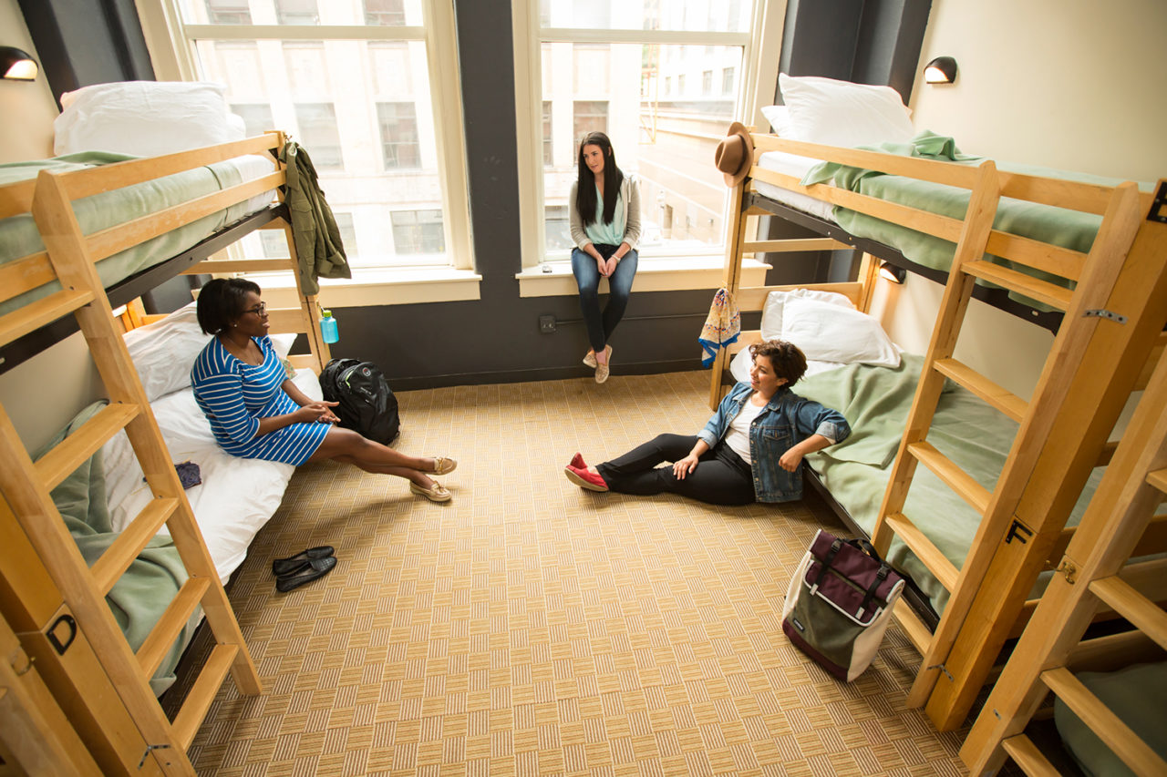 Guests at HI Chicago hostel sit chatting in a dorm room with large windows and several sets of freshly made bunk beds