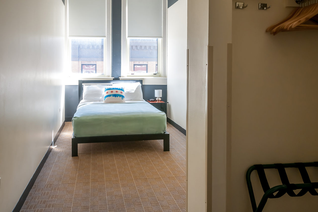 a private room at HI Chicago hostel featuring one freshly made full-sized bed, a closet with luggage rack, and two large windows.
