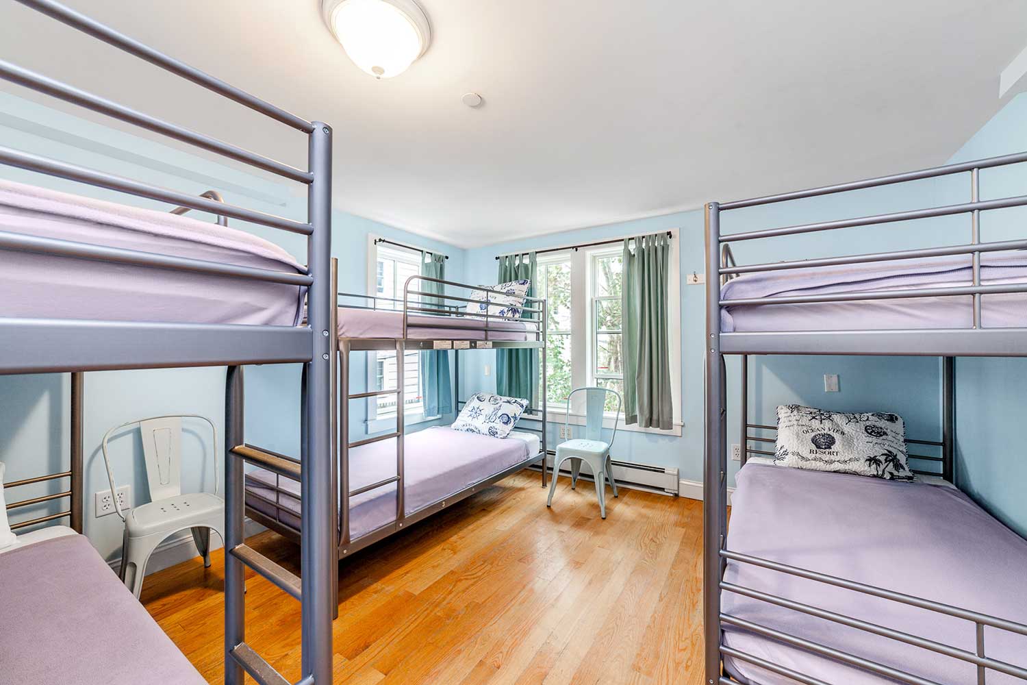 a bright shared dorm room at HI Hyannis hostel, with three sets of freshly made twin-sized bunk beds, warm hardwood flooring, and light blue walls.