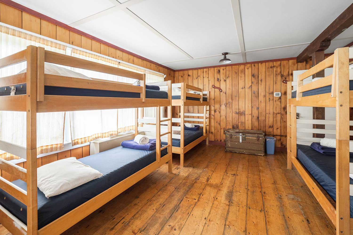 At HI Martha's Vineyard hostel, three sets of twin-sized bunk beds in a dorm room with hardwood floors and wood paneling on the walls. There is sunlight coming in through the windows.