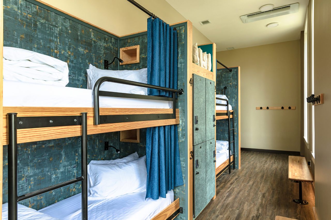A dorm room at HI New Orleans hostel with two sets of twin-sized bunk beds. Each bed has a privacy curtain, and secure lockers for guests' belongings separate each set of bunks for added privacy. The beds are made up with crisp white linens and each has a built-in reading light at the head.