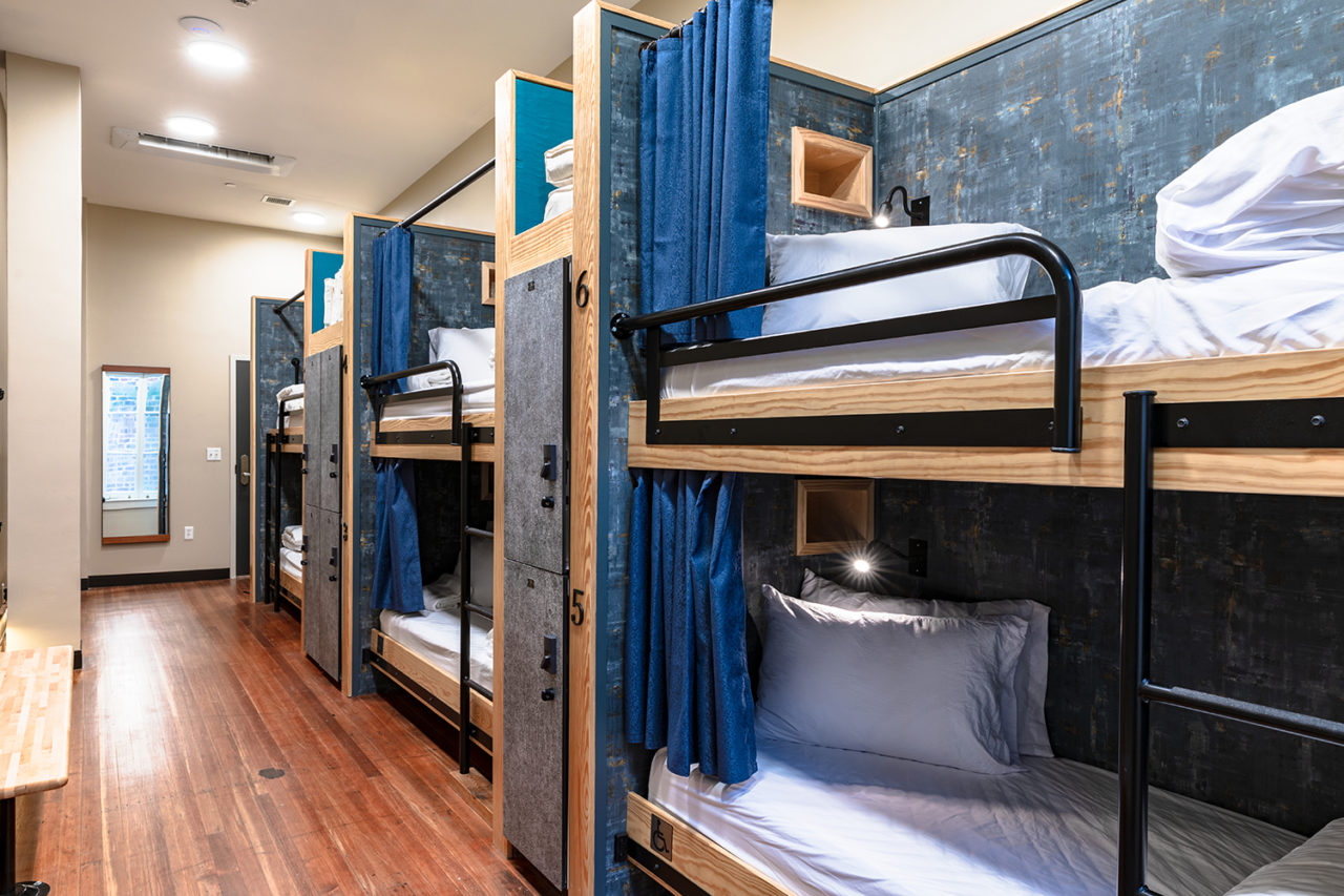 A dorm room at HI New Orleans hostel with three sets of twin-sized bunk beds. Each bed has a privacy curtain, and secure lockers for guests' belongings separate each set of bunks for added privacy. The beds are made up with crisp white linens and each has a built-in reading light at the head.