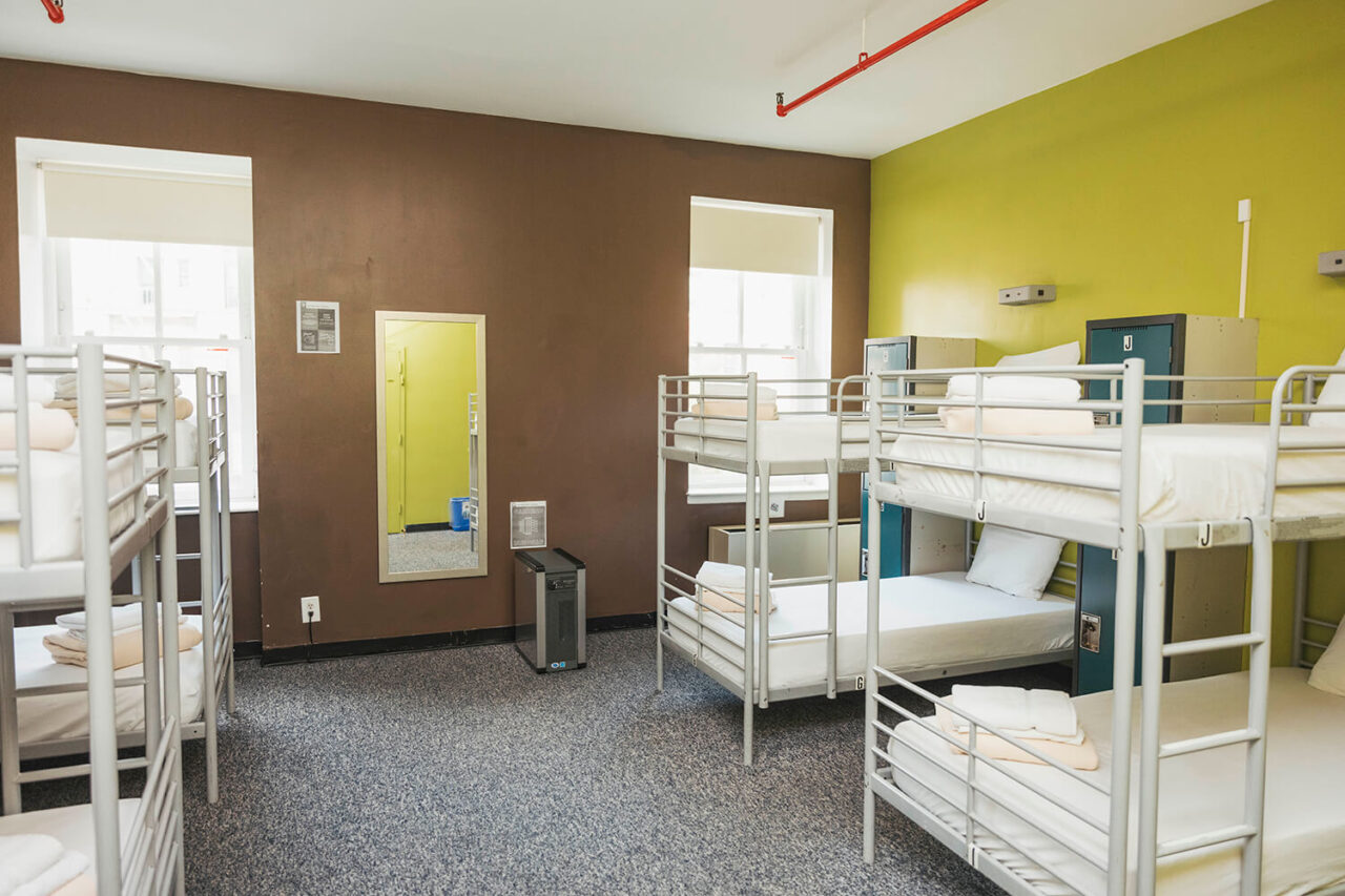A dorm room at HI New York City hostel with multiple sets of bunk beds with crisp white linens, secure lockers for guest belongings, and carpeted floors.