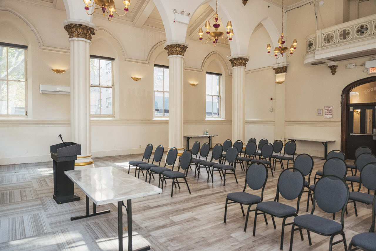 a large room with high ceilings and columns, set up with folding chairs facing a podium. The space is available to rent by groups staying at HI New York City hostel as well as outside groups.