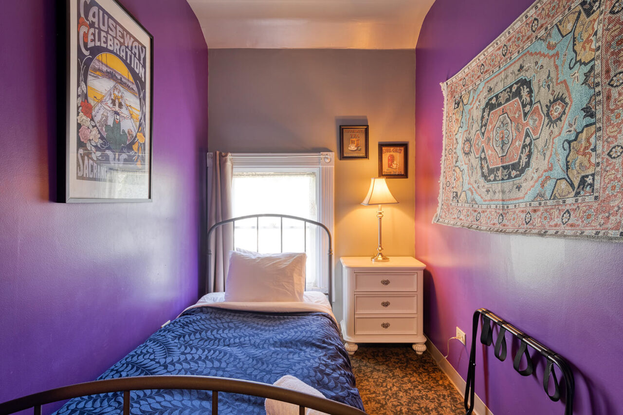 a cozy private single room at HI Sacramento hostel with one twin-sized bed, purple walls, and a window letting in natural light