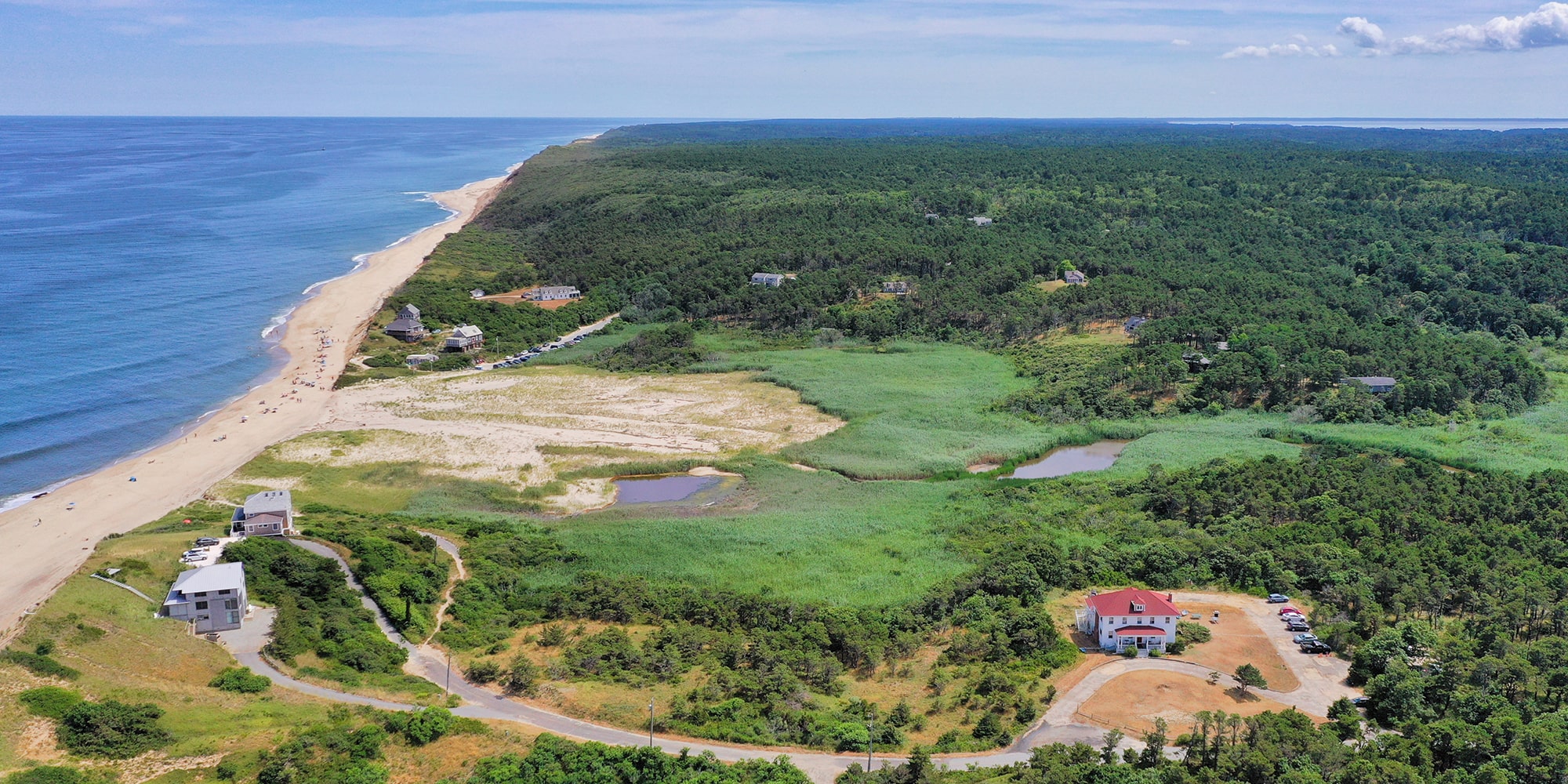 An aerial view of HI Truro hostel on Cape Cod. The red roof of the hostel is visible alongside a path leading to a beautiful beach.