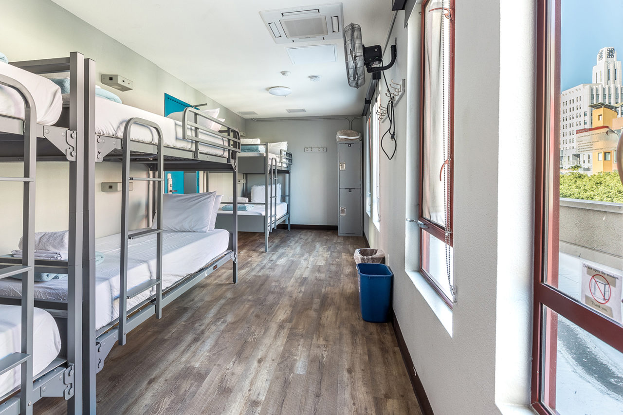 a long and narrow dorm room at HI Los Angeles Santa Monica with three sets of freshly made twin-sized bunk beds and two large windows overlooking the city.