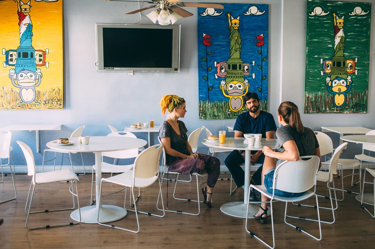 Three guests sit talking at a table over free breakfast at HI San Diego Downtown hostel. They are sitting at white tables on white chairs, and there are colorful paintings hanging on the wall behind them.