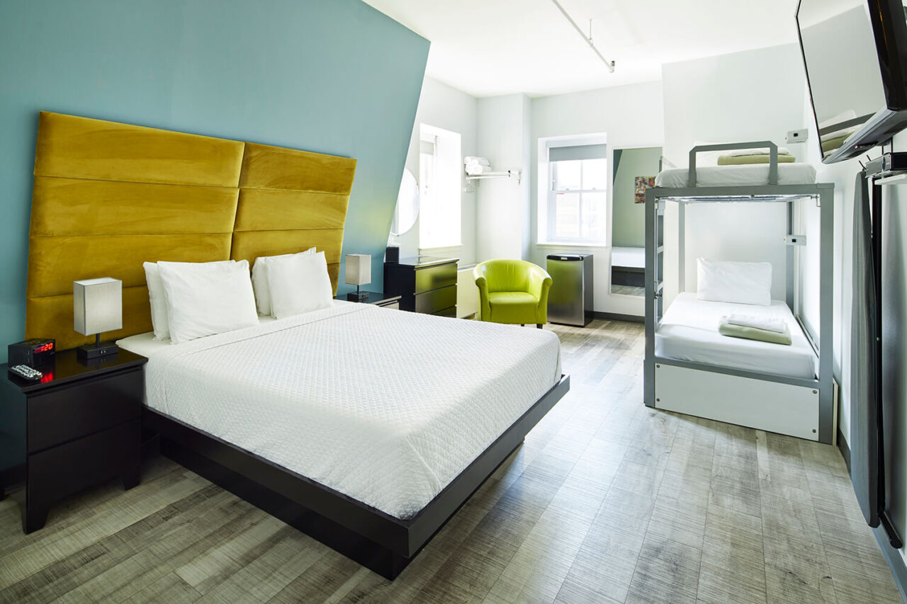 a private room at HI New york City hostel containing a queen-sized bed with a yellow velvet headboard, one set of twin-sized bunk beds, a sitting area with armchair, and a minifridge