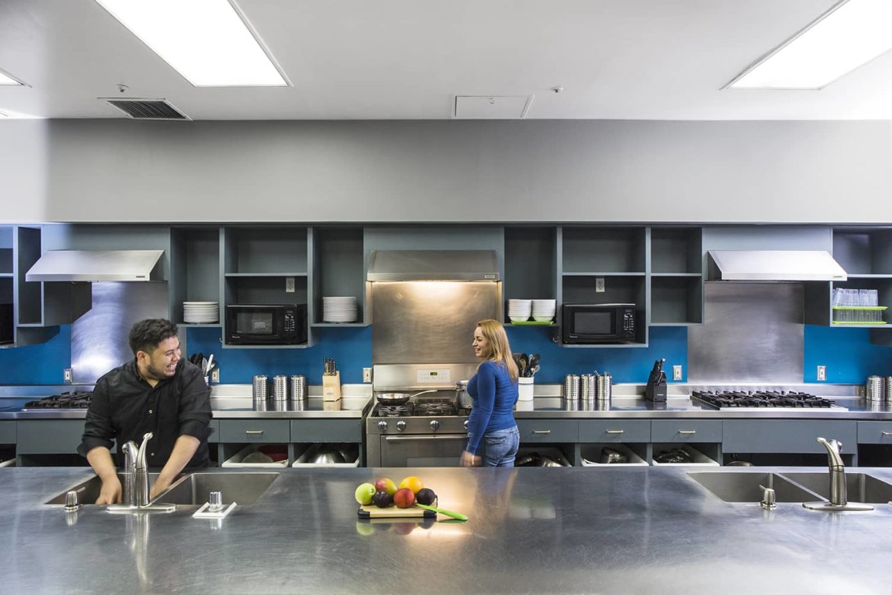 two people do dishes and cook on a stovetop in a modern stainless steel kitchen at HI Los Angeles Santa Monica hostel. The guest kitchen comes equipped with stoves, refrigerators, pots and pans, and everything else you need to prepare your own meals on site.