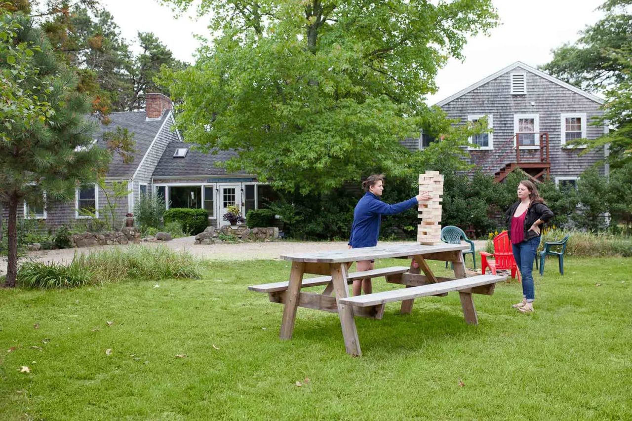 two guests play giant jenga on top of a picnic table on a large green lawn outside HI Martha's Vineyard hostel