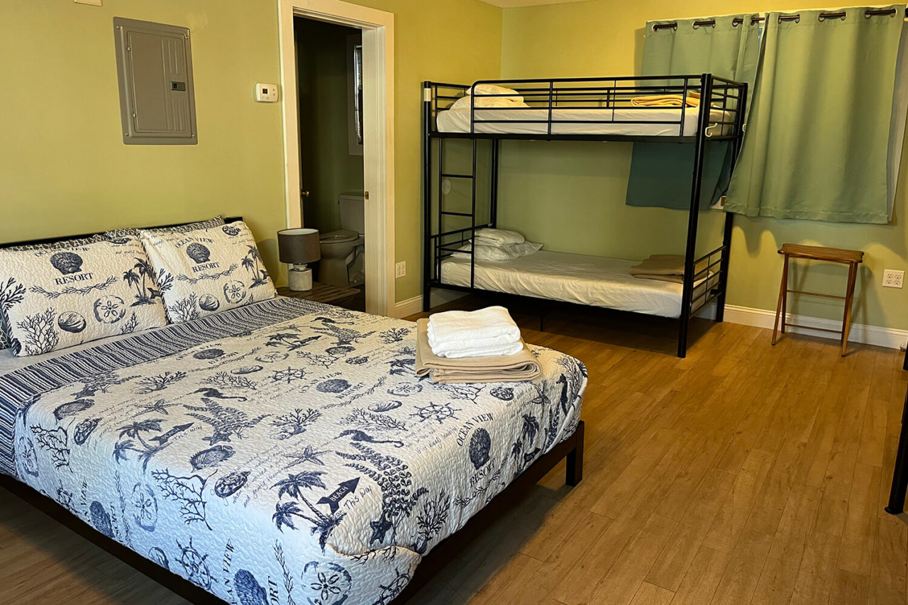a queen-sized bed and one set of twin-sized bunk beds inside a private cottage at HI Hyannis hostel. In the background, an interior doorway opens up onto a private ensuite bathroom.